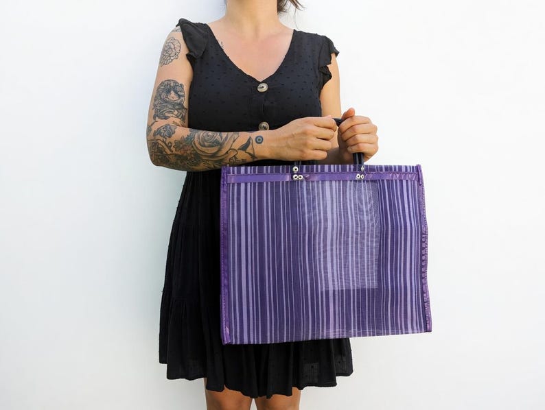 May include: A rectangular tote bag made of a semi-transparent purple mesh material featuring vertical stripes in varying shades of purple and white. The bag has a reinforced top border with silver-toned metal rivets and two sturdy dark handles.