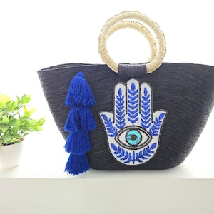 Könnte beinhalten: Schwarze Tragetasche mit gewebtem Design, mit einer aufgenähten Hamsa-Hand mit blauem Auge und blauen Akzenten. Die Tasche hat einen Strohgriff und eine blaue Quaste. Links ist eine kleine Topfpflanze.