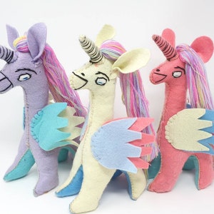 Puede incluir: Tres peluches de unicornio hechos a mano en fieltro, en colores lavanda, crema y rosa. Cada criatura presenta un cuerno en espiral, ojos bordados, crines de hilo multicolor y alas de fieltro superpuestas en tonos azul, rosa y crema.