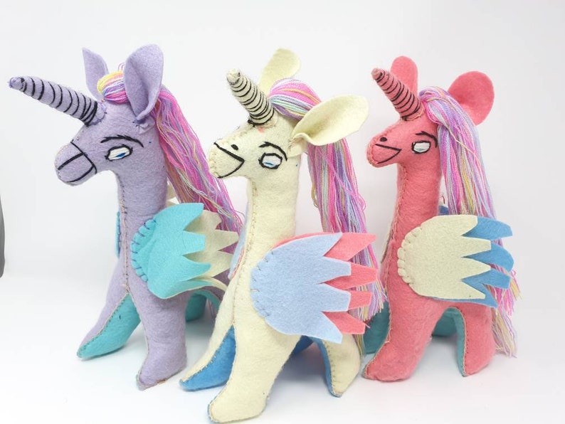 Puede incluir: Tres peluches de unicornio hechos a mano en fieltro, en colores lavanda, crema y rosa. Cada criatura presenta un cuerno en espiral, ojos bordados, crines de hilo multicolor y alas de fieltro superpuestas en tonos azul, rosa y crema.