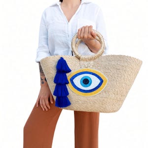 Puede incluir: Un bolso tipo tote de paja tejida con un gran motivo de ojo turco bordado en tonos azul, blanco y amarillo. El bolso incluye dos asas redondas y rígidas de paja y una borla decorativa de varias capas en color azul real en el lateral.