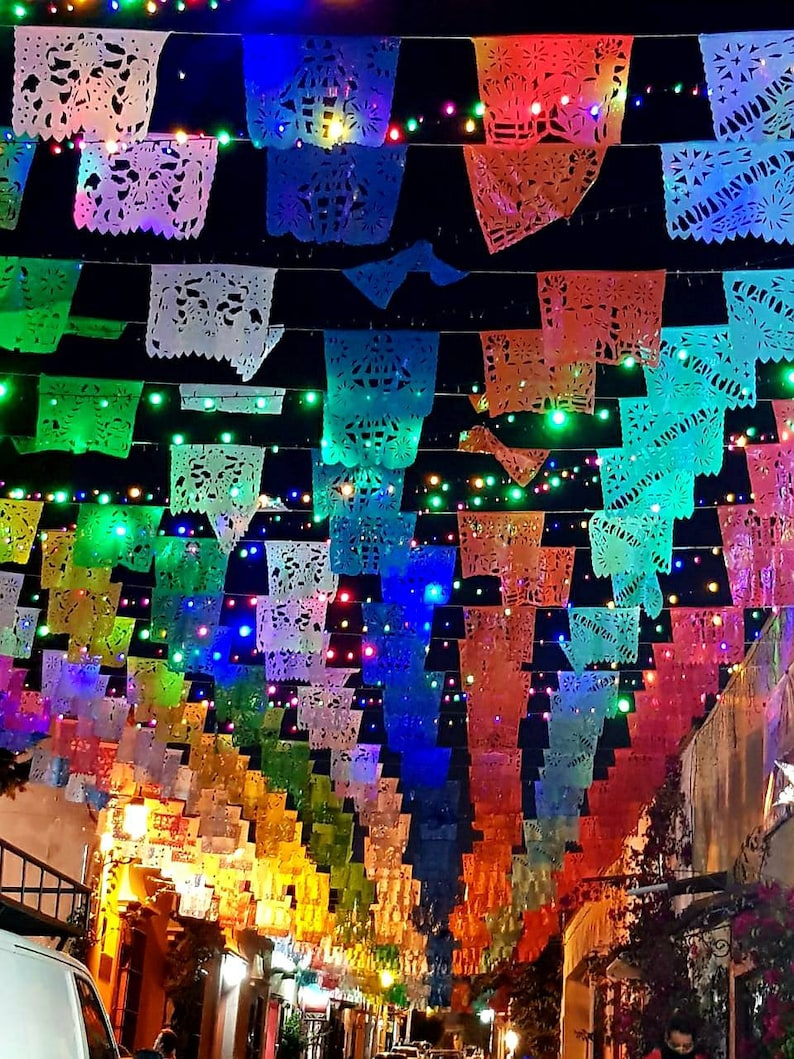May include: A street decorated with colorful paper lanterns, known as "papel picado", strung across the street. The lanterns are illuminated by twinkling lights, creating a festive atmosphere.