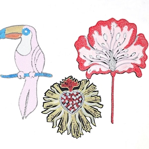 May include: Three embroidered patches featuring a pink toucan, a red flower, and a gold heart with rays. The toucan has a blue beak and black eyes. The flower has a pink center and red petals. The heart is surrounded by gold rays.