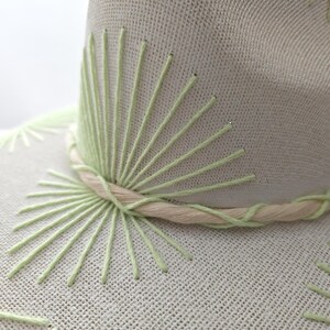 May include: A light beige hat featuring light green embroidered sunburst patterns on the crown and brim. A twisted beige and light green cord wraps around the base of the crown, accentuating the textured fabric of the hat's construction.