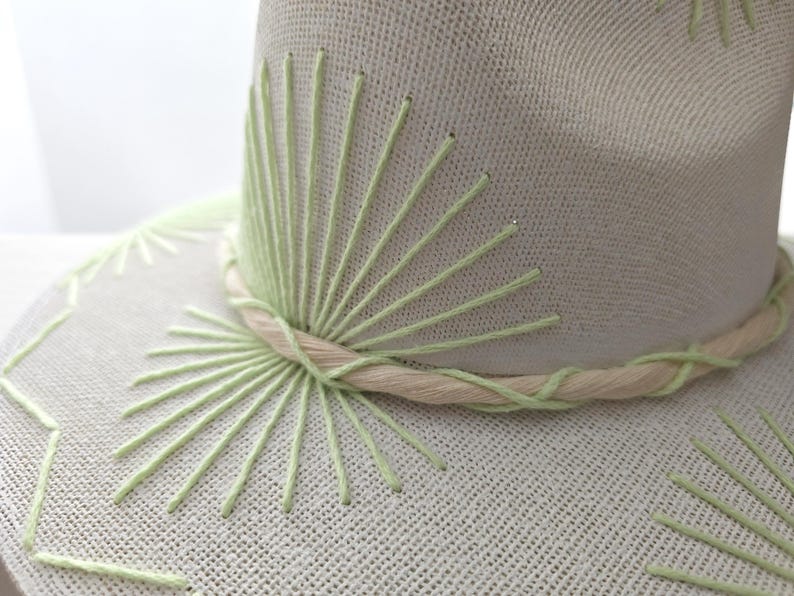 May include: A light beige hat featuring light green embroidered sunburst patterns on the crown and brim. A twisted beige and light green cord wraps around the base of the crown, accentuating the textured fabric of the hat's construction.