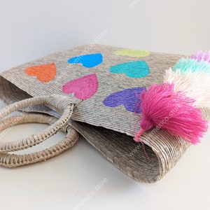 May include: A woven straw tote bag featuring two round handles and a series of painted hearts in orange, blue, pink, purple, and yellow. A cluster of colorful yarn tassels in shades of pink, white, and light blue hangs from the side of the bag.