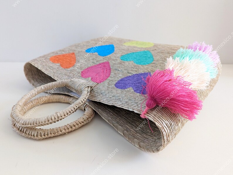 May include: A woven straw tote bag featuring two round handles and a series of painted hearts in orange, blue, pink, purple, and yellow. A cluster of colorful yarn tassels in shades of pink, white, and light blue hangs from the side of the bag.