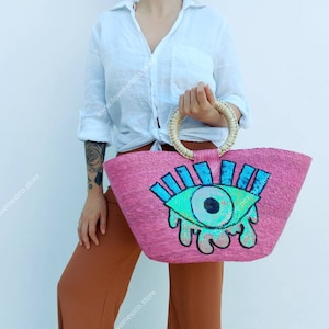May include: Pink straw tote bag with a large blue and green eye design. The eye has sequins and a white pupil. The bag has a woven handle.
