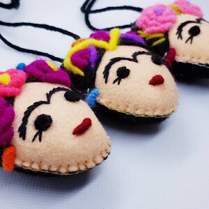 May include: Three handmade felt pendants featuring stylized faces with embroidered black eyebrows, red lips, and colorful floral headpieces. Each pendant is attached to a black cord, showcasing detailed stitching and vibrant yarn accents in pink, yellow, and purple.