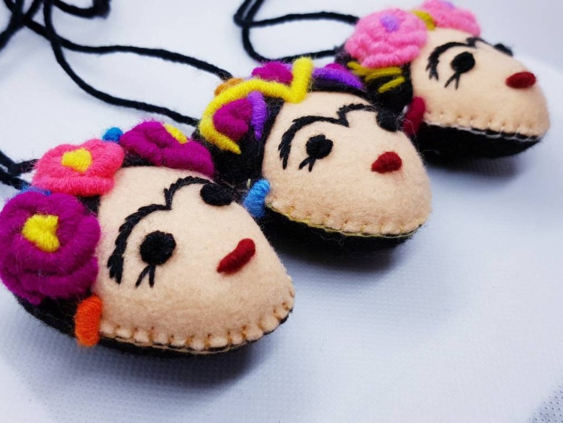 May include: Three handmade felt pendants featuring stylized faces with embroidered black eyebrows, red lips, and colorful floral headpieces. Each pendant is attached to a black cord, showcasing detailed stitching and vibrant yarn accents in pink, yellow, and purple.