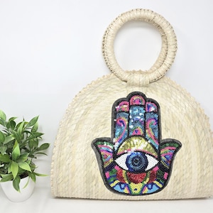 以下が含まれることがあります： 丸いハンドルとカラフルなハmsaの手と邪眼のデザインが施されたストローのトートバッグ。バッグは自然な織りストローで作られており、薄いベージュの色をしています。