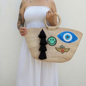 Puede incluir: Un bolso de paja trenzada con asas redondas, que presenta un gran parche de lentejuelas de ojo azul y blanco, un parche de cara sonriente verde, un parche de corazón alado dorado y una larga borla negra colgando del asa.