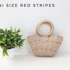 May include: A woven straw handbag with a round handle and red stripes, featuring the text MINI SIZE RED STRIPES. The bag has a tapered shape and a textured natural fiber construction with small red horizontal accents throughout the exterior.