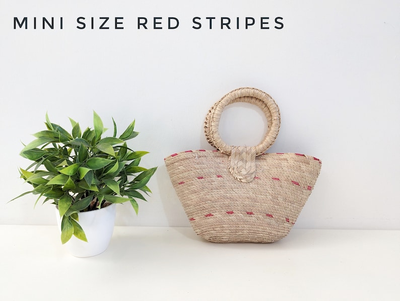 May include: A woven straw handbag with a round handle and red stripes, featuring the text MINI SIZE RED STRIPES. The bag has a tapered shape and a textured natural fiber construction with small red horizontal accents throughout the exterior.