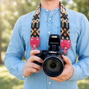 May include: A black Nikon DSLR camera with a lens attached, suspended by a camera strap featuring a woven geometric pattern in shades of brown, black, and cream, finished with bright pink leather ends and silver metal rivets.