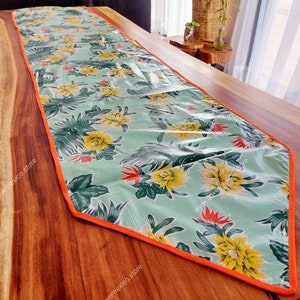 May include: A green and yellow floral table runner with an orange border. The runner is made of a vinyl-like material and has a tropical print.