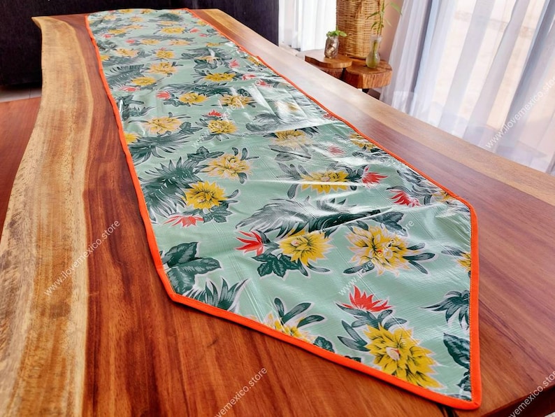 May include: A green and yellow floral table runner with an orange border. The runner is made of a vinyl-like material and has a tropical print.