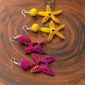 Puede incluir: Dos pares de pendientes colgantes con gancho plateado. Un par consta de esferas envueltas en hilo amarillo con dijes en forma de estrella de mar. El segundo par presenta esferas envueltas en hilo magenta y amarillo con dijes de mariposa.