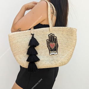 以下が含まれることがあります： 黒いたッセルと刺繍されたハートのついた手のモチーフのパッチが付いたベージュのトートバッグ。パッチは赤、ピンク、金色のディテールと白い真珠のアクセントが施されています。