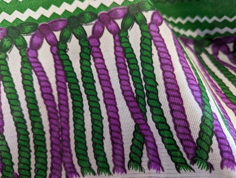 May include: A fabric featuring a repeating pattern of vertical, twisted rope-like strands in alternating shades of deep purple and forest green against a white background, topped with a horizontal band of green and white zigzag chevron stripes.