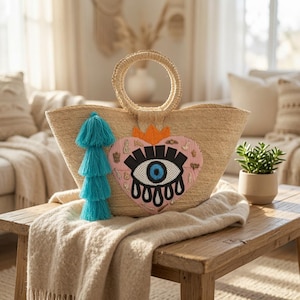 Op de afbeelding: Een gevlochten strooien tas met twee ronde handvatten, voorzien van een grote roze hartvormige patch met een geborduurd zwart en blauw oogontwerp, een oranje kroondetail en een lange, meerlaagse fel turquoise kwast aan de zijkant.