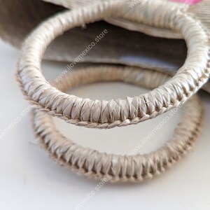 May include: Two circular bag handles crafted from light beige woven natural fibers. The handles feature a tightly coiled, braided texture with a consistent thickness throughout their circumference, attached to a larger woven bag body.