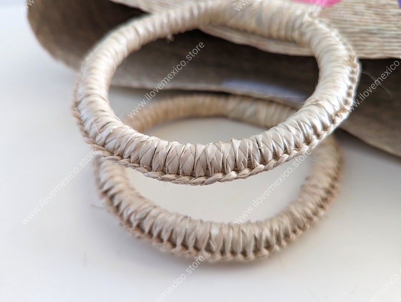 May include: Two circular bag handles crafted from light beige woven natural fibers. The handles feature a tightly coiled, braided texture with a consistent thickness throughout their circumference, attached to a larger woven bag body.