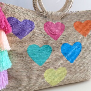 May include: A woven straw tote bag featuring six painted hearts in purple, pink, orange, teal, blue, and yellow. A large tassel made of four stacked layers in bright pink, peach, mint green, and light purple hangs from the side of the bag.