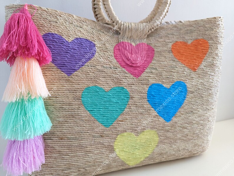 May include: A woven straw tote bag featuring six painted hearts in purple, pink, orange, teal, blue, and yellow. A large tassel made of four stacked layers in bright pink, peach, mint green, and light purple hangs from the side of the bag.