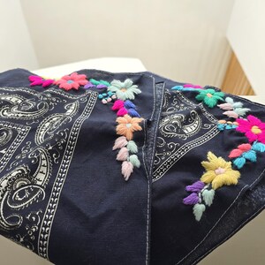 May include: A dark navy blue bandana featuring a traditional white paisley print on one side and vibrant, multi-colored floral embroidery along the edge. The embroidery includes bright pink, yellow, light blue, and soft pastel thread accents.