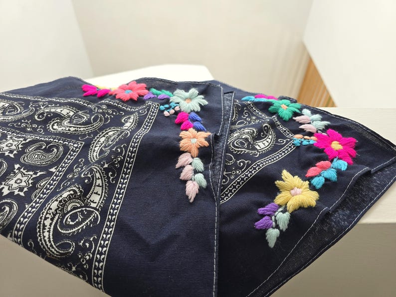 May include: A dark navy blue bandana featuring a traditional white paisley print on one side and vibrant, multi-colored floral embroidery along the edge. The embroidery includes bright pink, yellow, light blue, and soft pastel thread accents.