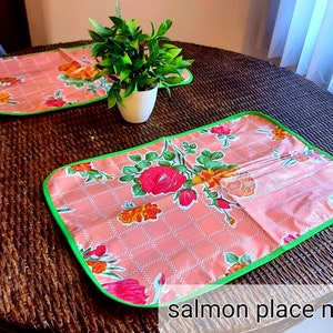 May include: Two salmon-colored placemats with a green border and floral pattern. The placemats are on a brown wicker table.