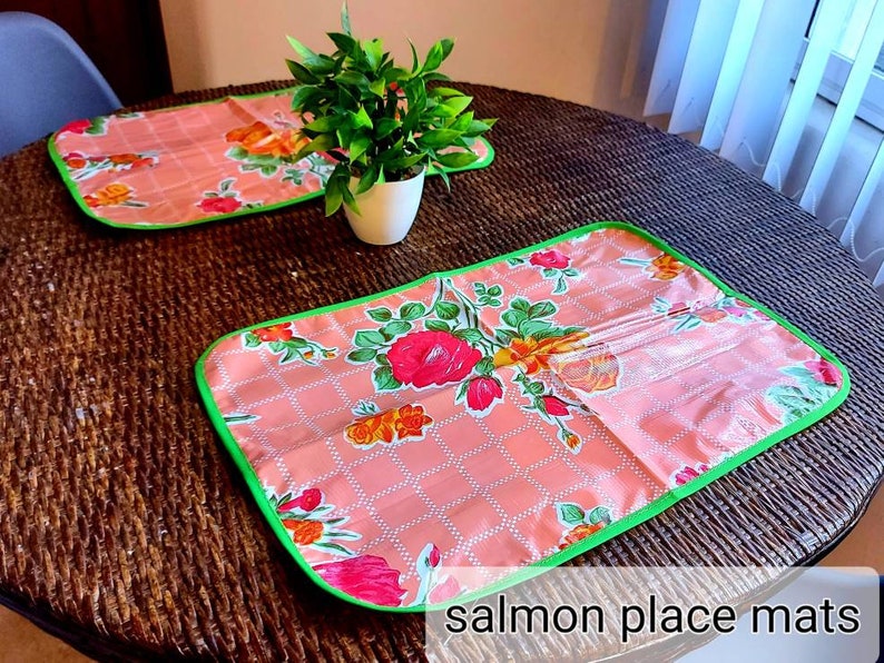 May include: Two salmon-colored placemats with a green border and floral pattern. The placemats are on a brown wicker table.