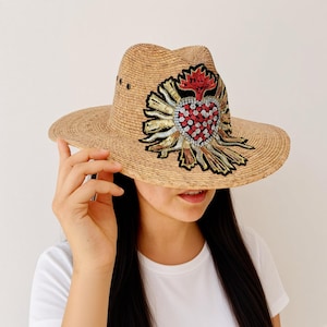 May include: A tan straw fedora hat featuring a large, decorative patch on the front. The patch displays a stylized heart design embellished with red, silver, and gold sequins, surrounded by radiating rays in black, gold, and silver thread embroidery.