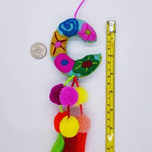 May include: A blue felt letter C with colourful embroidered flowers and a pink, yellow and green pom-pom trim. The letter is approximately 10 cm tall.