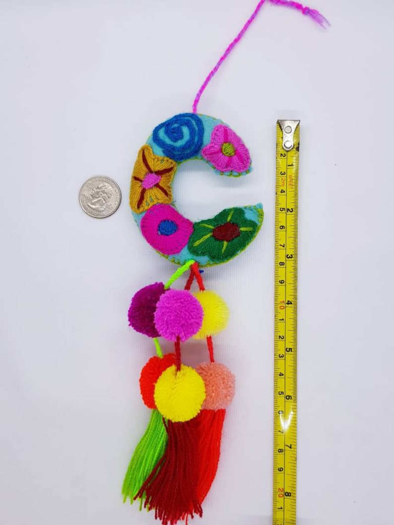 May include: A blue felt letter C with colourful embroidered flowers and a pink, yellow and green pom-pom trim. The letter is approximately 10 cm tall.