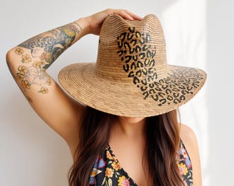 Puede incluir: Un sombrero fedora de paja color canela con un patrón de leopardo negro que se extiende desde la copa hasta el ala. El sombrero tiene una copa pellizcada y un ala ancha y estructurada, mostrando una textura tejida natural en todo el material.