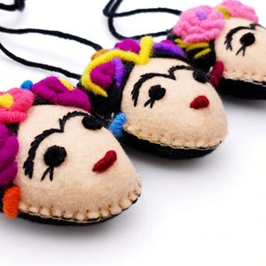 May include: Three felt pendants on black cords, each shaped like a face with embroidered black eyebrows, eyes, and red lips. Each pendant features colorful felt flowers in shades of pink, purple, and yellow attached to the top of the head.