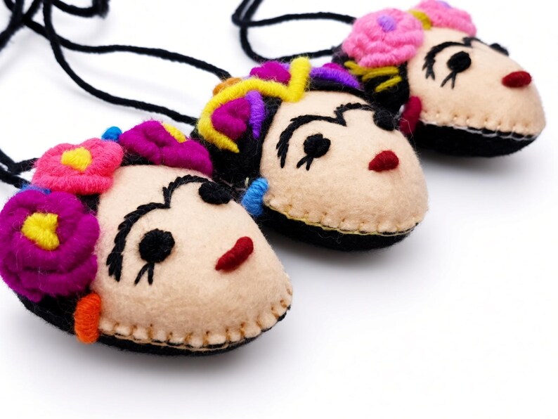 May include: Three felt pendants on black cords, each shaped like a face with embroidered black eyebrows, eyes, and red lips. Each pendant features colorful felt flowers in shades of pink, purple, and yellow attached to the top of the head.