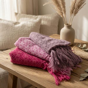 May include: A stack of three folded woven blankets in shades of magenta, light pink, and mauve, each featuring a repeating geometric diamond pattern and long, loose fringe along the edges, resting on a wooden surface with a textured appearance.