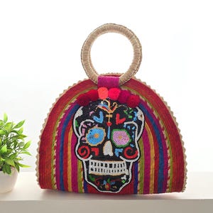 Puede incluir: Bolso tote colorido hecho a mano con un diseño de calavera del Día de Muertos. El bolso tiene un cuerpo tejido con rayas rojas, azules y amarillas, un asa redonda y pompones decorativos. La calavera está adornada con lentejuelas.