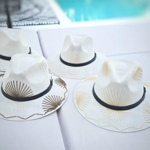 May include: Four white fedora hats with black bands. Two hats feature plain white crowns and brims, while the other two are decorated with brown and gold sunburst-style embroidery patterns on the brims and crowns, featuring scalloped edges.