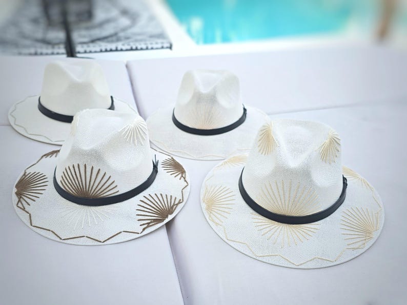 May include: Four white fedora hats with black bands. Two hats feature plain white crowns and brims, while the other two are decorated with brown and gold sunburst-style embroidery patterns on the brims and crowns, featuring scalloped edges.