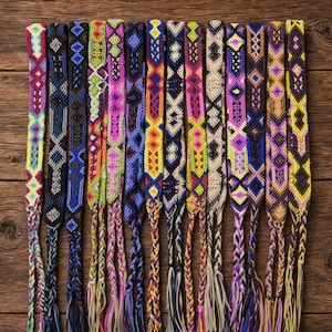 Op de afbeelding: Een collectie van twaalf geweven vriendschapsarmbandjes met ingewikkelde geometrische patronen in levendige kleuren zoals roze, blauw, geel, paars, oranje en zwart. Elk armbandje eindigt in een gevlochten koord met losse draadkwastjes.