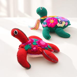 Könnte beinhalten: Zwei Schildkröten-Stofftiere aus Filz. Eine Schildkröte ist rot mit bunter Blumenstickerei und sichtbaren Nähten an den Flossen. Die andere ist grün mit ähnlicher Blumenstickerei und einer mehrfarbigen Garnquaste um den Hals.