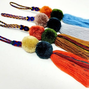 May include: Five colourful pom-pom keychains with tassels. The pom-poms are in shades of orange, green, brown, blue, and yellow. The tassels are in various colours, including red, orange, brown, green, blue, and white.