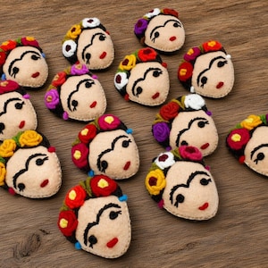 May include: A collection of twelve felted wool appliques, each shaped as a face with dark eyebrows, red lips, and a crown of colorful flowers in shades of red, yellow, purple, and white, arranged on a textured brown wooden surface.