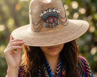 Puede incluir: Un sombrero fedora de paja color canela con un parche decorativo en forma de ojo hecho de cuentas, lentejuelas y cristales transparentes. El parche incluye cuentas colgantes en forma de lágrima azul oscuro y finas cadenas negras.