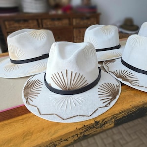 May include: A collection of white fedora hats featuring a woven texture, a black band around the crown, and decorative brown thread embroidery in a sunburst pattern on the crown and brim, with a zigzag stitch detail along the edge of the brim.