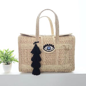 Peut inclure: Sac fourre-tout en paille tressée de forme rectangulaire avec fermeture à rabat. Le sac présente un motif d'œil décoratif avec des perles bleues et blanches et un pompon noir. Le sac a deux poignées.